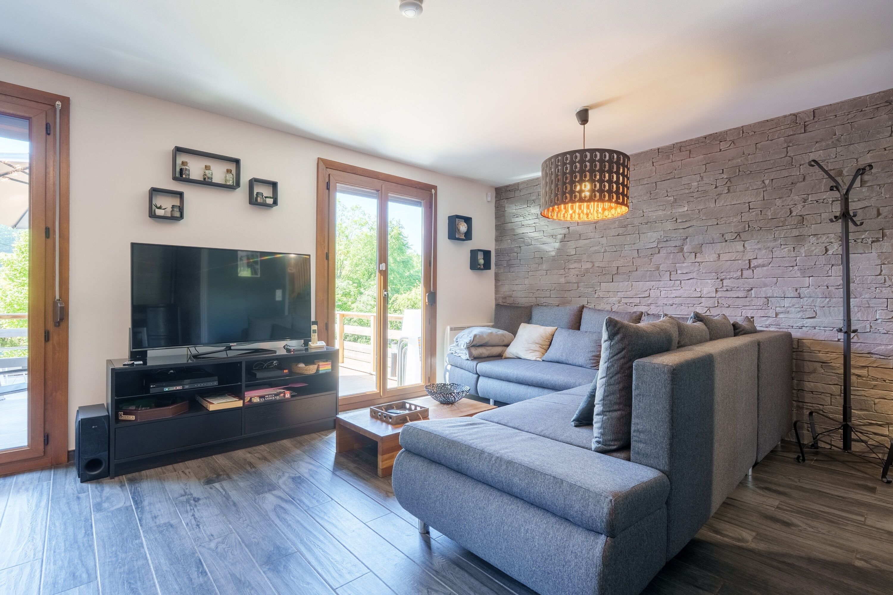 Una sala de estar moderna con televisión de pantalla plana, sofá gris, suelo de madera y pared de ladrillo.