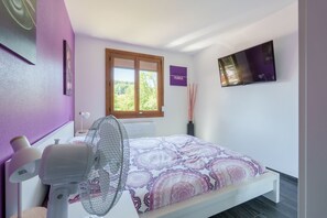 Ein Schlafzimmer mit einem Bett, einem Standventilator, einem Fenster mit Jalousien, einem an der Wand befestigten Fernseher und einem violetten Schild.
