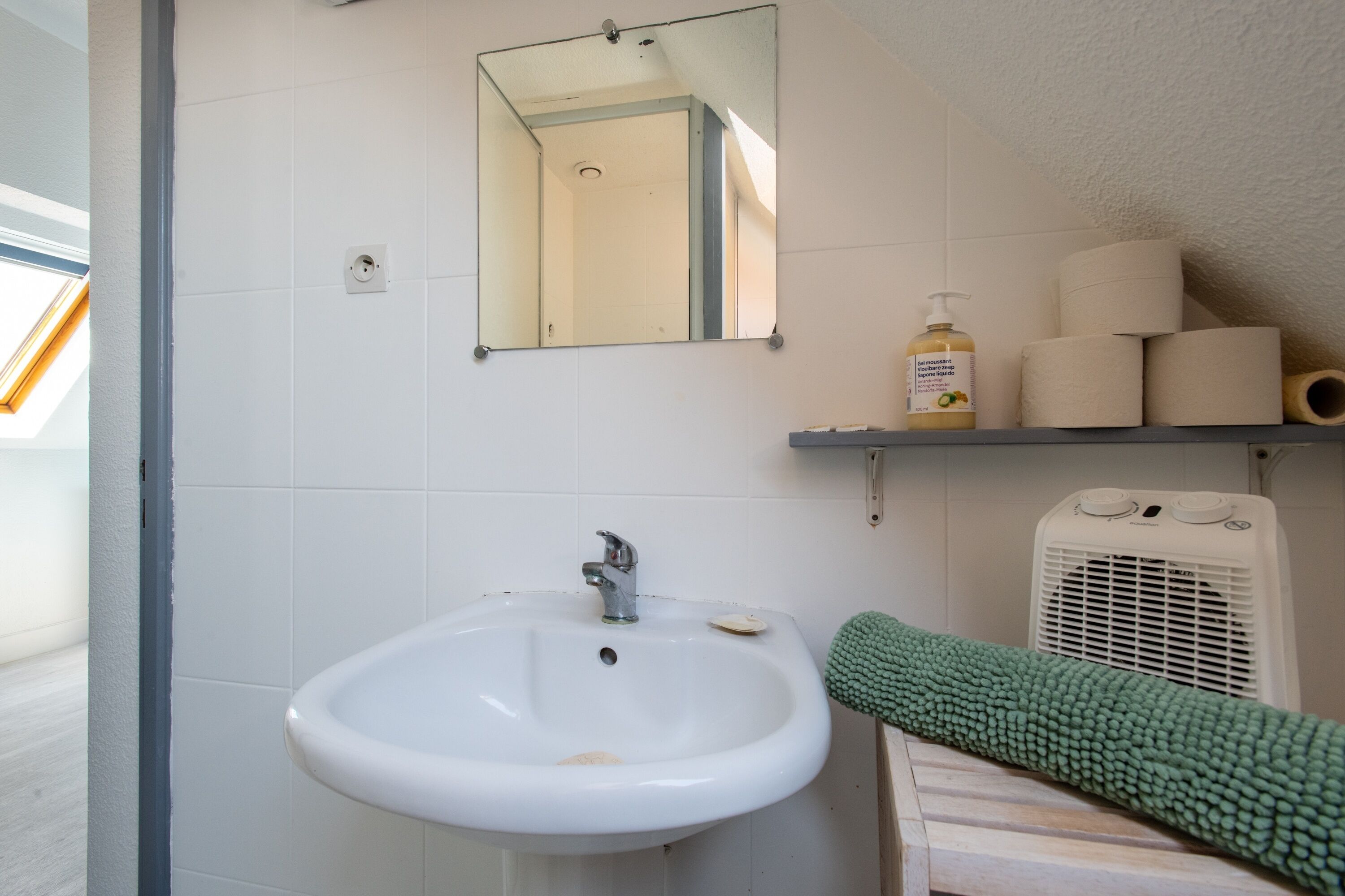 A bathroom with a white sink, a mirror, a towel rack with towels, a bottle of liquid soap, and a fan.