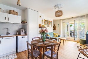 A compact kitchen with a sink, coffee machine, and white cabinets. There is a dining area with a round table, chairs, and a vase with flowers. The room has a large window with a view of the outdoors. - Azur & Magnolia - À 750 m de la plage (La Seyne-sur-Mer)