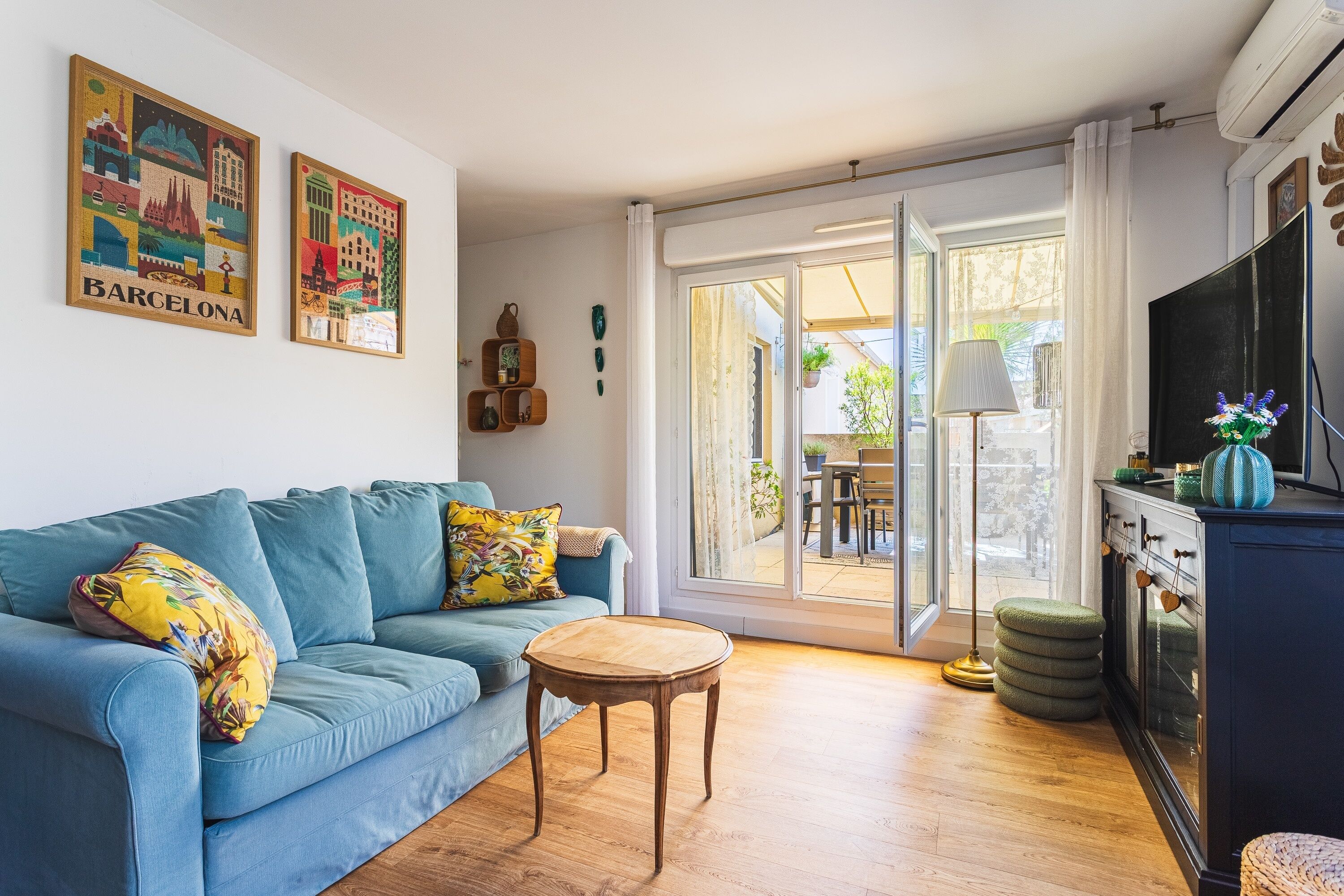 Un salon avec un canapé bleu, une petite table en bois et un téléviseur posé sur un meuble. Deux tableaux ornent le mur, et une porte-fenêtre coulissante donne accès à un espace extérieur.