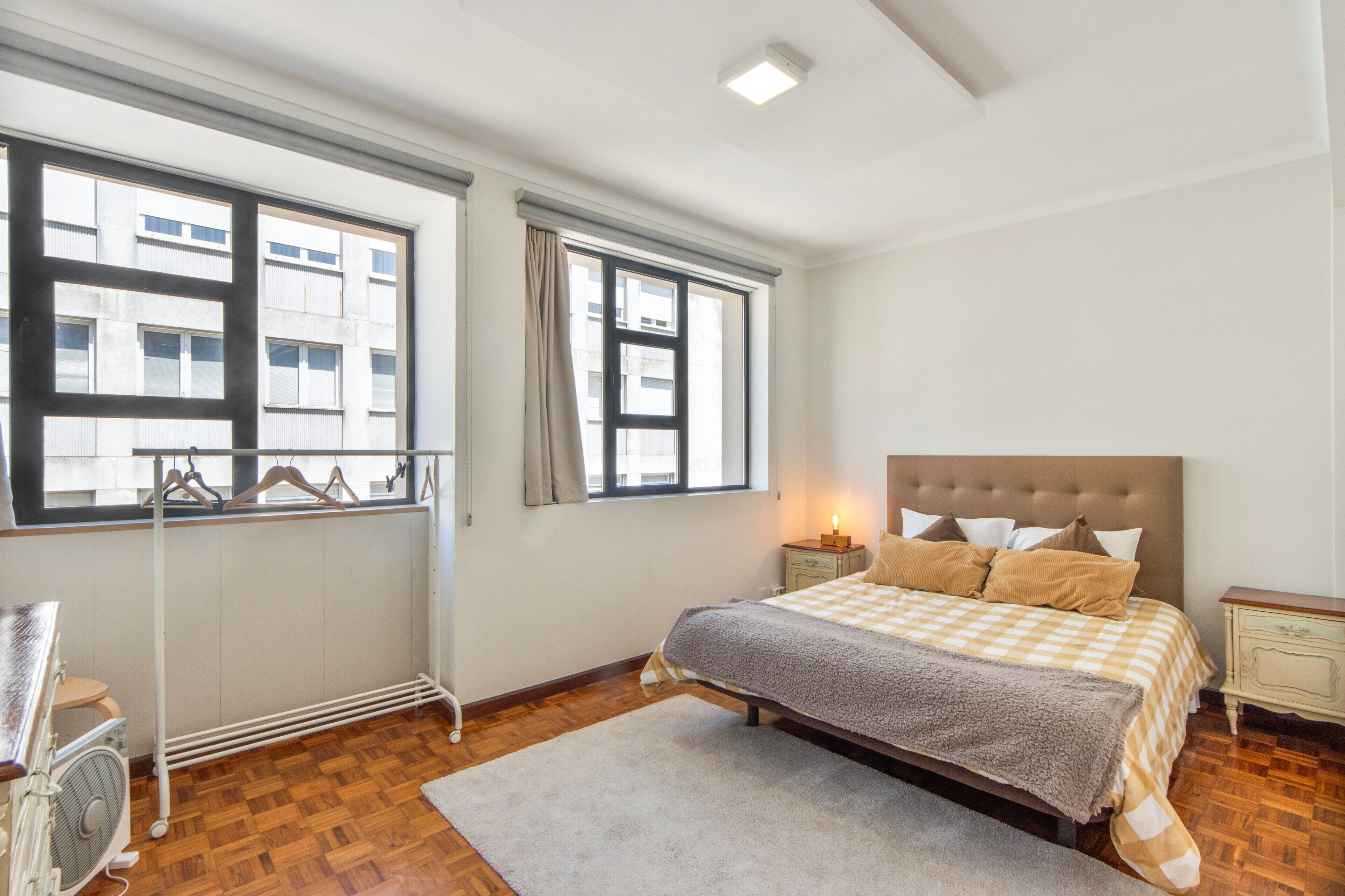 A bedroom with a bed, a radiator, a window with a view of buildings, and a bedside table with a lamp.