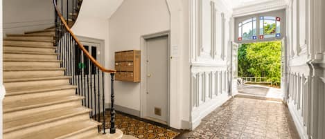 A grand staircase with a wooden handrail, a patterned floor, and a view of a lush garden through an open door.