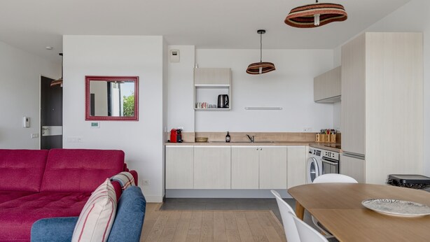 A modern kitchen with white cabinets, a wooden dining table, and a red-framed mirror. - Les Perles de l’Aiguillon - Appt face au bassin (La Teste-de-Buch)