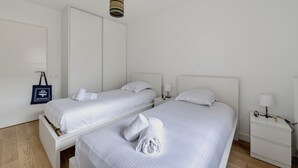 A hotel room with two beds, a nightstand with a lamp, a wall-mounted towel rack, and a hanging light fixture. - Les Perles de l’Aiguillon - Appt face au bassin (La Teste-de-Buch)