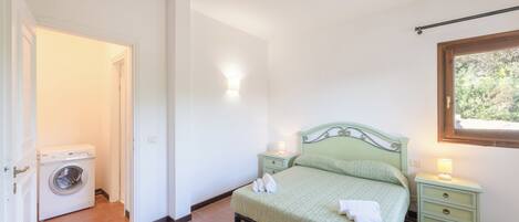 A bedroom with a bed, a nightstand, a wall-mounted light, a window, and a washing machine in the adjacent room.