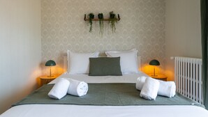 A neatly made bed with white linens, two green pillows, and two rolled towels. There are two bedside lamps, a wall-mounted shelf with plants, and a radiator. - L'Alma du Laudon - Centre village et climatisation (Saint-Jorioz)