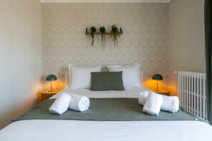 A neatly made bed with white linens, two green pillows, and two rolled towels. There are two bedside lamps, a wall-mounted shelf with plants, and a radiator. - L'Alma du Laudon - Centre village et climatisation (Saint-Jorioz)