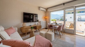A modern living room with a sofa, a TV, a dining area, and a view of the mountains through large windows. - L'Alma du Laudon - Centre village et climatisation (Saint-Jorioz)