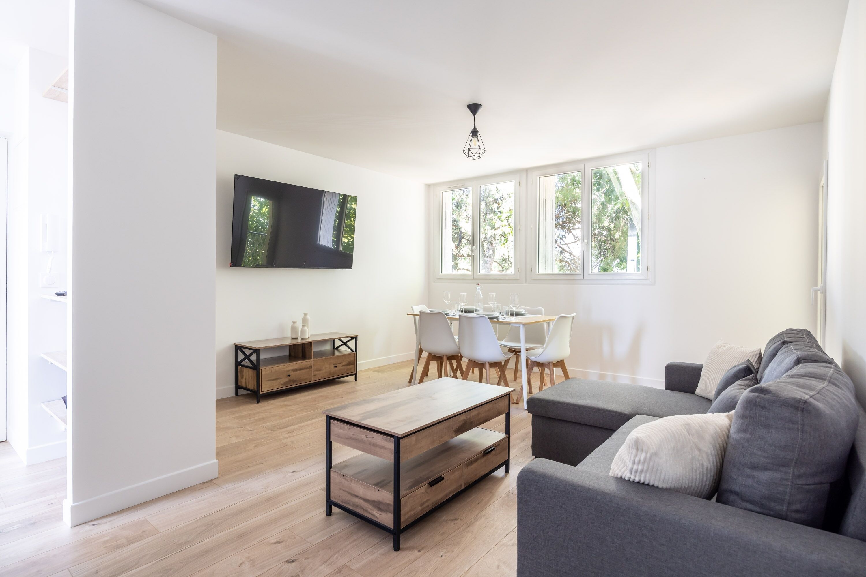 Una sala de estar moderna con un sofá gris, una mesa de centro de madera, un televisor montado en la pared y un comedor con mesa y sillas.