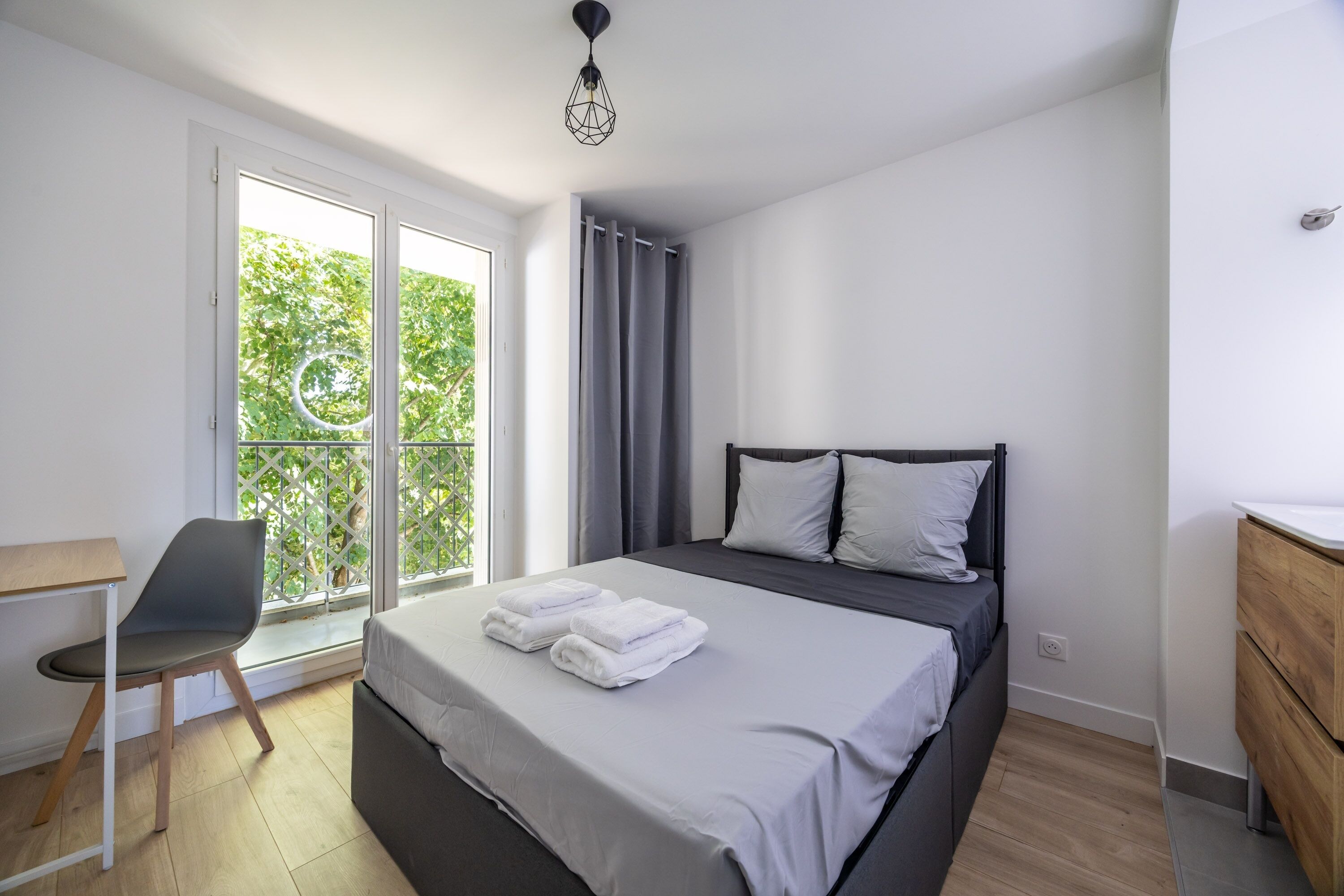 Un dormitorio con cama, escritorio, silla, ventana con cortinas y vista a follaje.