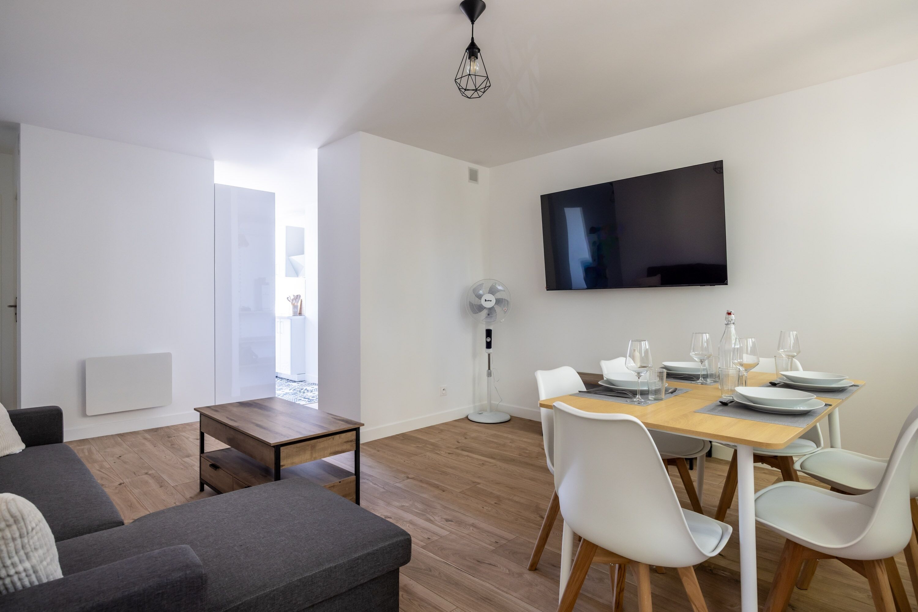 Una sala moderna con un sofá, una mesa de comedor preparada para cuatro personas, un televisor de pantalla plana y un ventilador de pie.