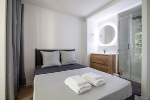 Quarto moderno com cama, penteadeira de madeira, espelho redondo e box de vidro.
