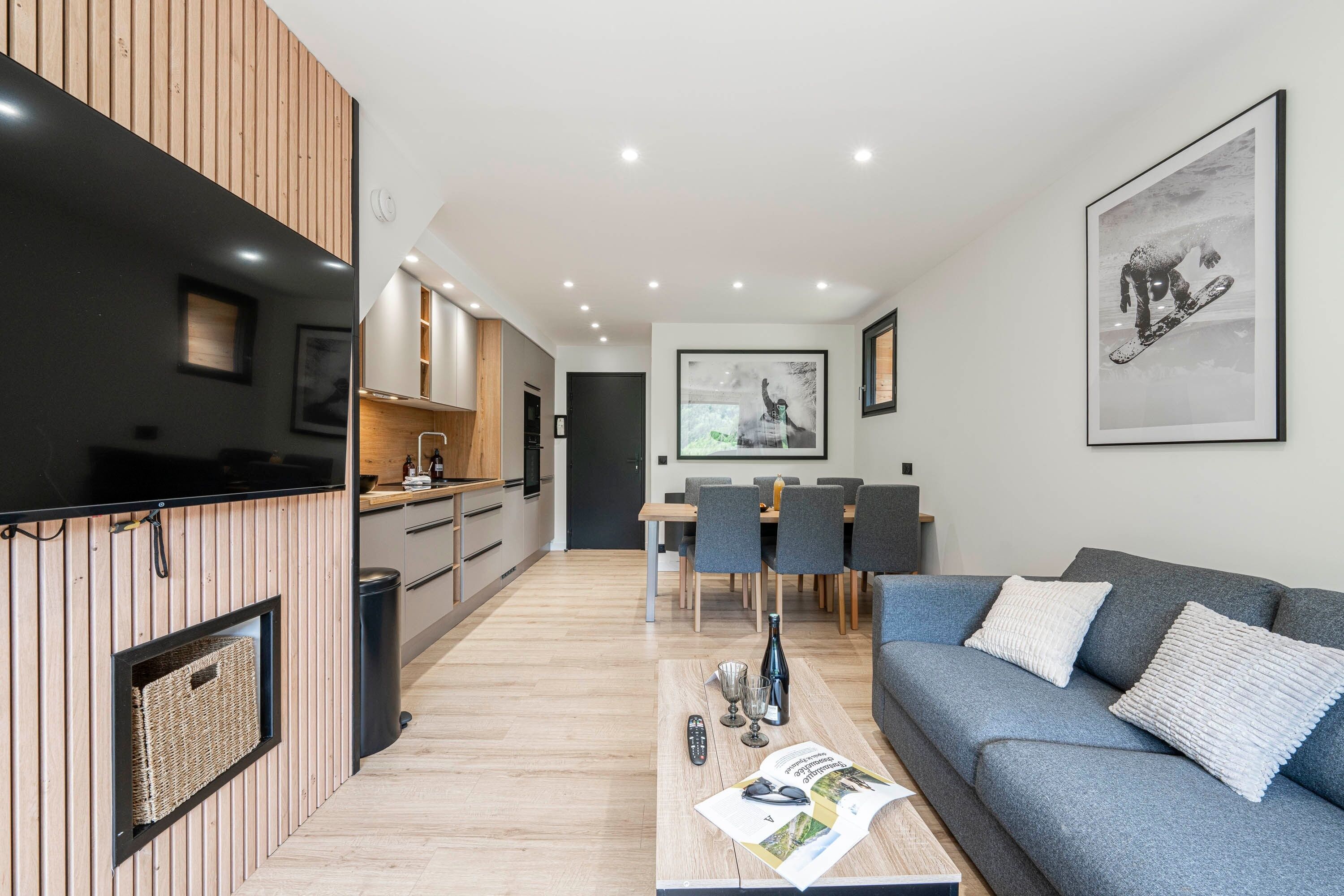A modern living room with a sofa, a coffee table with magazines, a TV, and a dining area with chairs and a table.