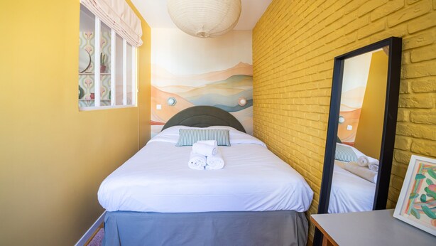 Una cama individual con sábanas blancas y almohada a rayas, una mesita de noche, un espejo y una pared con un mural.