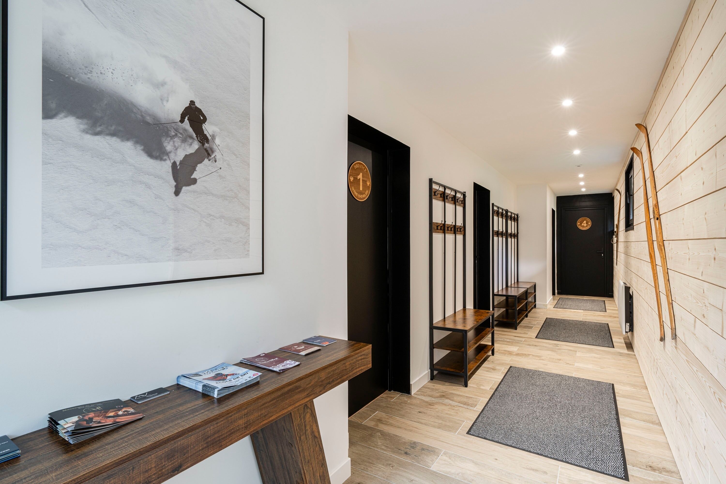 A hallway with a ski-themed artwork, a wooden desk with magazines, and a staircase with ski poles leaning against the wall.