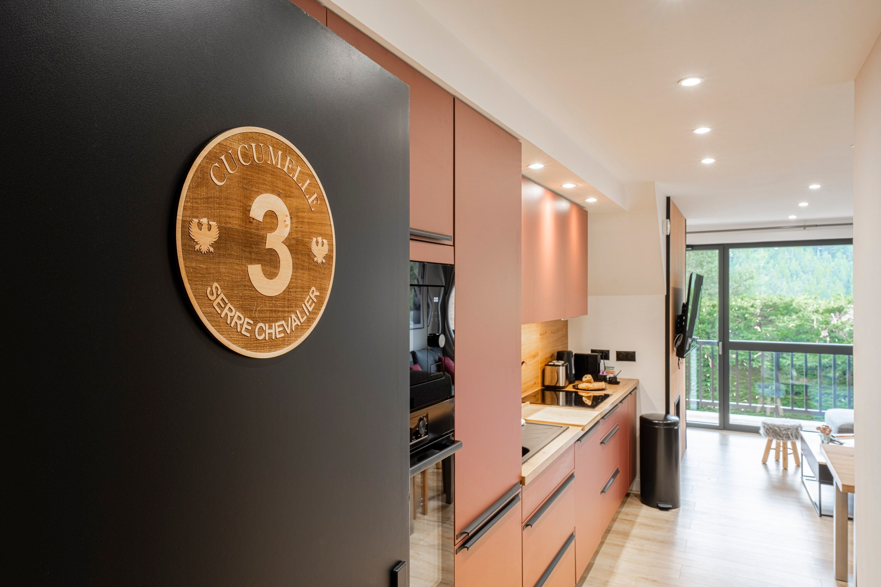 A modern kitchen with a large window, a black wall with a wooden plaque, and a view of greenery outside.
