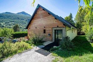 Una cabaña de madera con base de piedra, rodeada de abundante vegetación y vistas a la montaña.