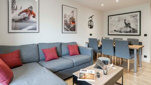 A modern living room with a grey sofa, red cushions, a coffee table with magazines, a laptop, and a dining area with grey chairs and a wooden table. - Suite Cucumelle - Beau Duplex pour 6 avec balcon (Saint-Chaffrey)
