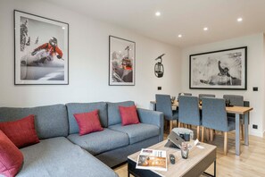 Ein modernes Wohnzimmer mit einem grauen Sofa, roten Kissen, einem Couchtisch mit Zeitschriften, einem Laptop und einem Essbereich mit grauen Stühlen und einem Holztisch.