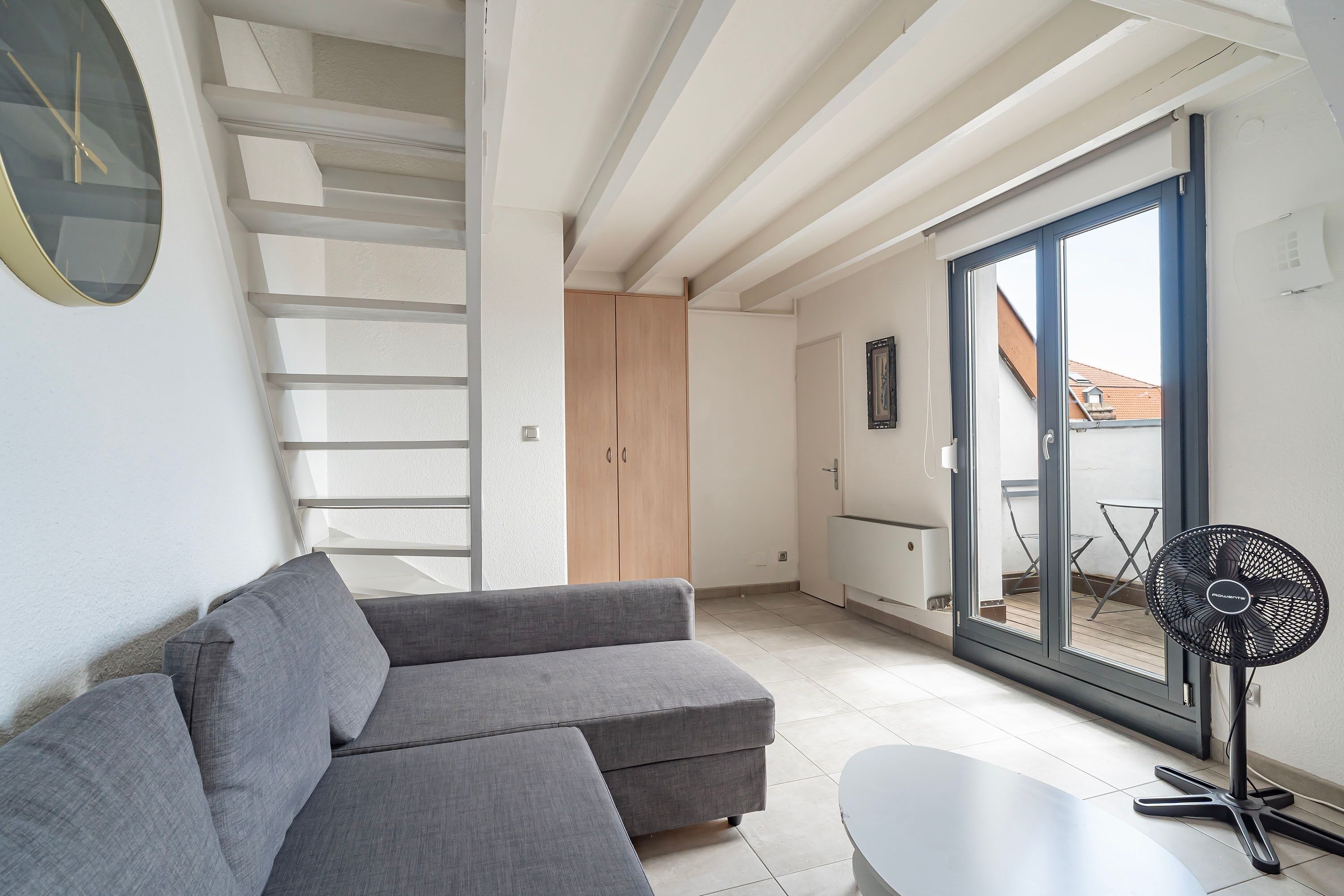 A modern living space with a grey sofa, a small table, a standing fan, a staircase, and a sliding glass door leading to an outdoor area.