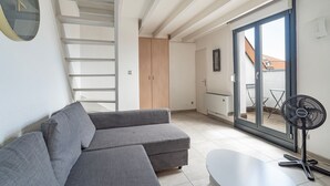 A modern living space with a grey sofa, a small table, a standing fan, a staircase, and a sliding glass door leading to an outdoor area. - La Réserve de Fathala - À 500 m de la gare (Sarrebourg)