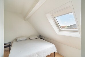 Ein Schlafzimmer mit schrägen Wänden, einem Bett mit weißer Bettwäsche, einem Nachttisch, einer Lampe und einem Fenster mit Blick auf Gebäude.