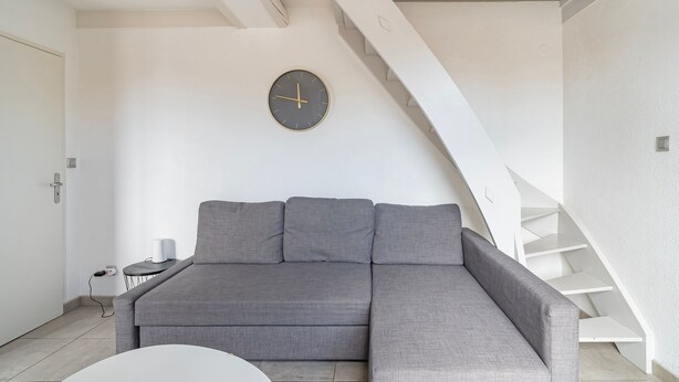A modern living room with a grey sectional sofa, a round wall clock, a white coffee table, and a staircase. - La Réserve de Fathala - À 500 m de la gare (Sarrebourg)