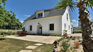 A white house with a grey roof, a patio with a table and chairs, and a palm tree in the foreground. - Fargesia - Jolie maison avec jardin, proche plage (Fréhel)