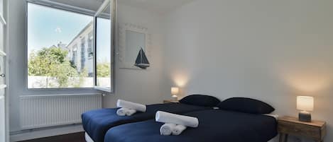 A bedroom with two beds, a window, a nightstand with a lamp, and a wall-mounted mirror.