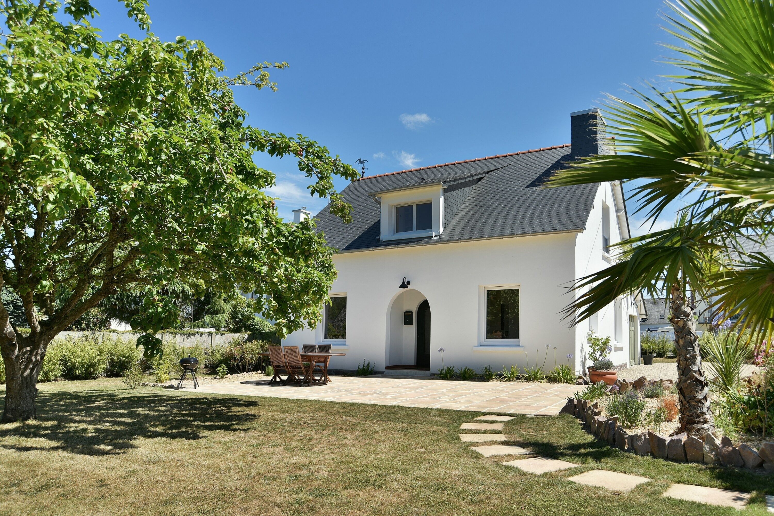 Una casa bianca con un patio piastrellato, un piccolo giardino e un albero sullo sfondo.