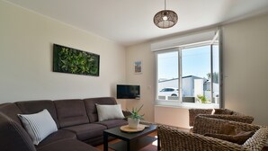 A living room with a brown sofa, a small table, a wicker chair, a hanging light fixture, and a window with a view of buildings. - Fargesia - Jolie maison avec jardin, proche plage (Fréhel)