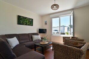 A living room with a brown sofa, a small table, a wicker chair, a hanging light fixture, and a window with a view of buildings. - Fargesia - Jolie maison avec jardin, proche plage (Fréhel)