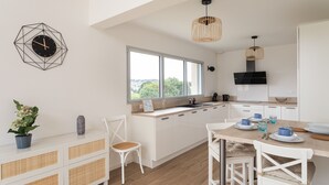 A modern kitchen with white cabinetry, a large window, and a dining area with a table set for two. - Les horizons - À moins d'1 km de la plage (Erquy)