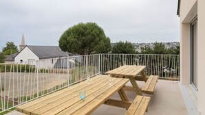 An outdoor patio with wooden tables and benches, a white fence, and a view of a church and trees. - Les horizons - À moins d'1 km de la plage (Erquy)