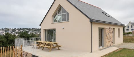 A single-story house with a pitched roof, a large window, and a wooden picnic table.