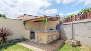 Ein Garten mit einer Holzdachterrasse, einer Bar mit Hockern, einer hängenden Pflanze und einem blauen Laternenleuchte.