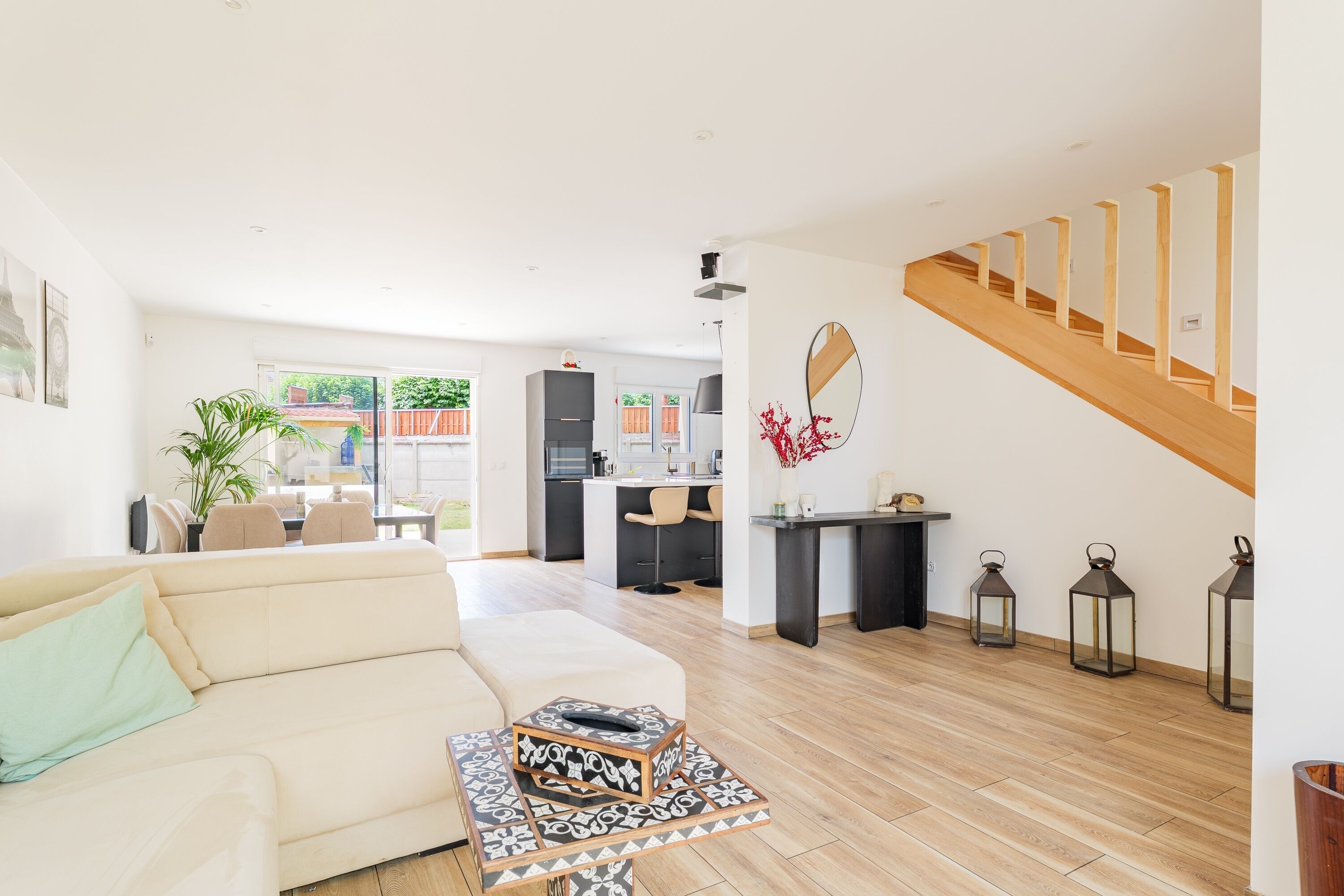 En moderne stue med sofa, sofabord og spiseplads med stole. Der er et køkken i baggrunden, en trappe og udsigt ud.