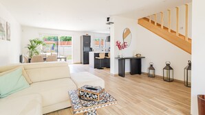 Ein modernes Wohnzimmer mit einer Couch, einem Couchtisch und einem Essbereich mit Stühlen. Im Hintergrund befindet sich eine Küche, eine Treppe und ein Blick ins Freie.
