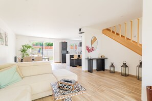 Ein modernes Wohnzimmer mit einer Couch, einem Couchtisch und einem Essbereich mit Stühlen. Im Hintergrund befindet sich eine Küche, eine Treppe und ein Blick ins Freie.