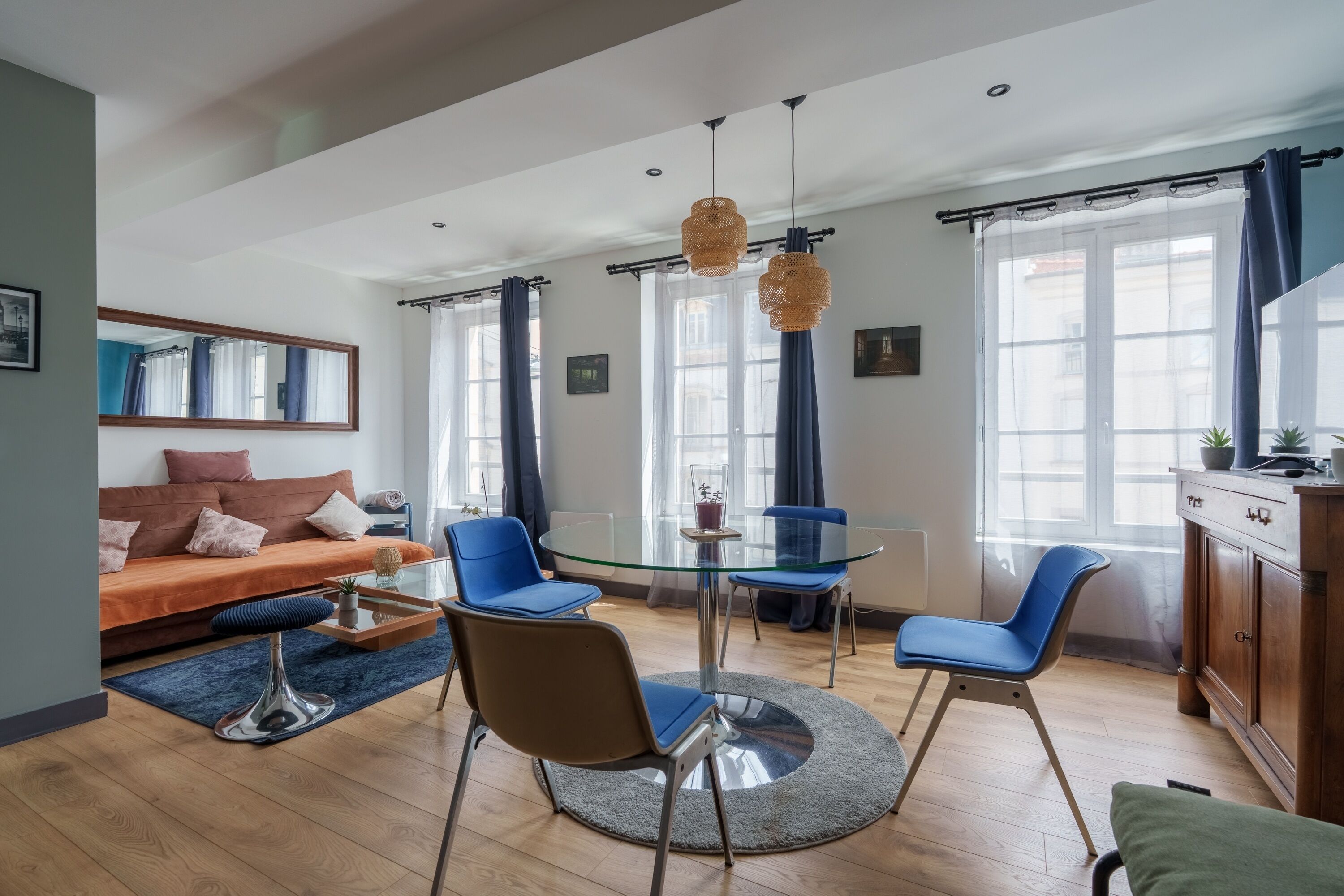 Un salon moderne avec une table à manger en verre, des chaises bleues, un canapé et une grande fenêtre avec des rideaux.