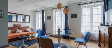 A modern living room with a glass dining table, blue chairs, a sofa, and a large window with curtains.