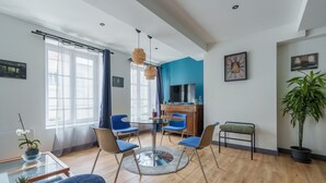 Ett modernt vardagsrum med ett soffbord i glas, blå fåtöljer, en rund matta, en tv och en stor krukväxt.