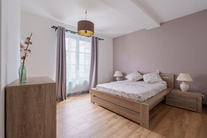 Ein Schlafzimmer mit einem hölzernen Bett, Nachttischen, einer Lampe, einem Fenster mit Vorhängen und einer Vase mit Trockenblumen.