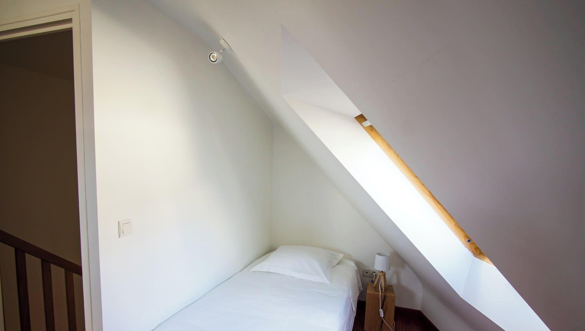 A small, single-bed room with a sloped ceiling, a wooden nightstand, and a wall-mounted light fixture.