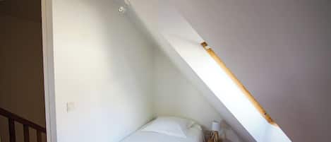 A small, single-bed room with a sloped ceiling, a wooden nightstand, and a wall-mounted light fixture.