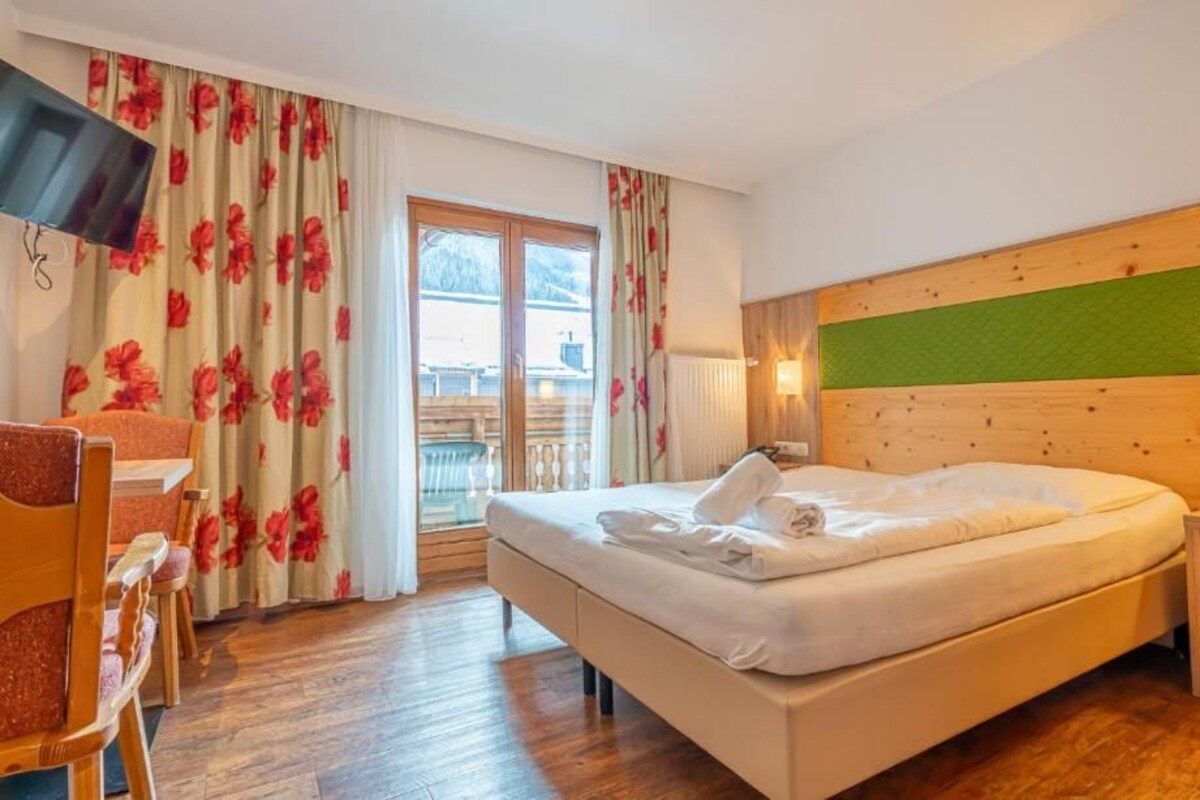 Una camera d'albergo con un letto, una scrivania e una sedia. Si vede una finestra con tende, un televisore a parete e una testiera di legno.