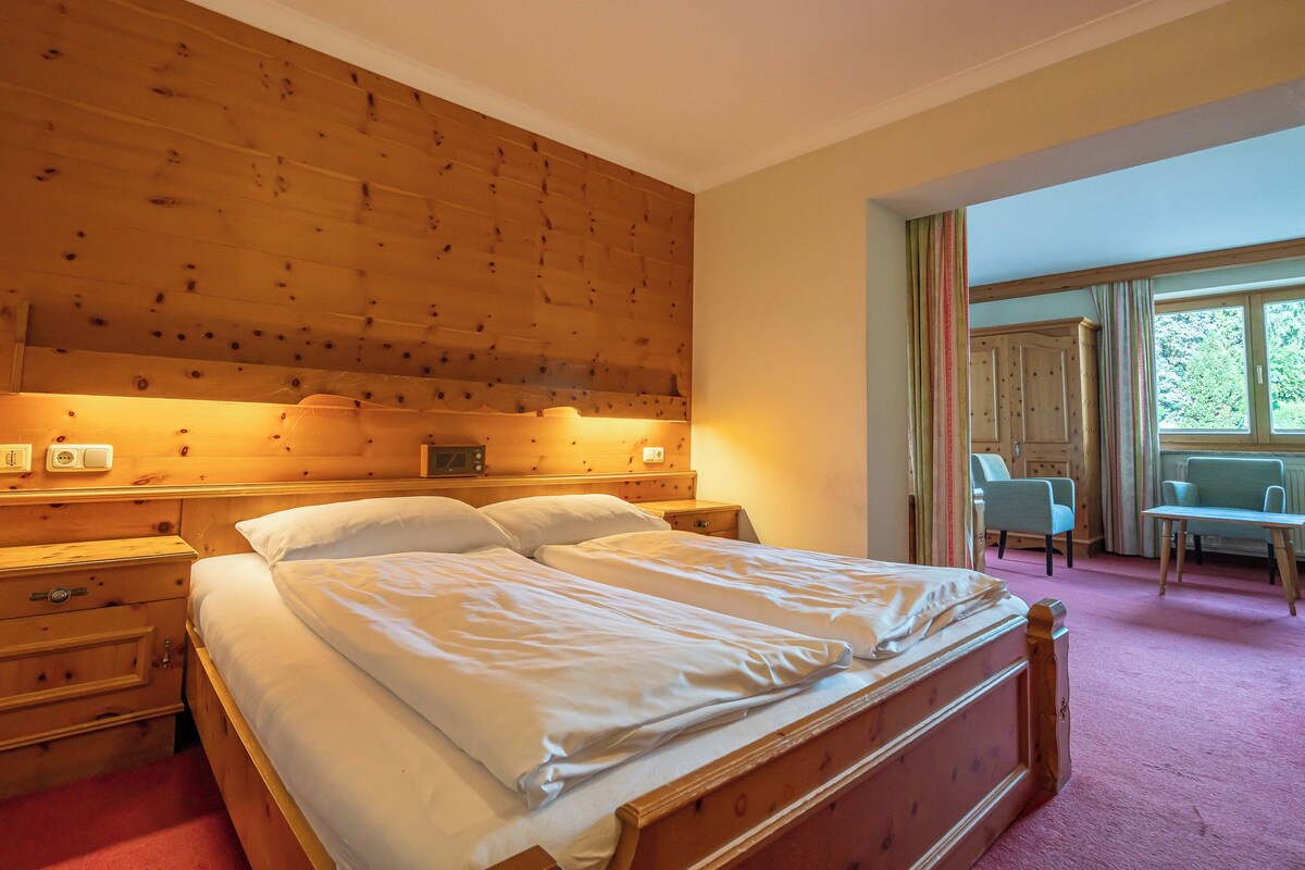 Una camera d'albergo con una testiera in legno, un letto con lenzuola bianche, un comodino con una radio e una sedia vicino alla finestra.