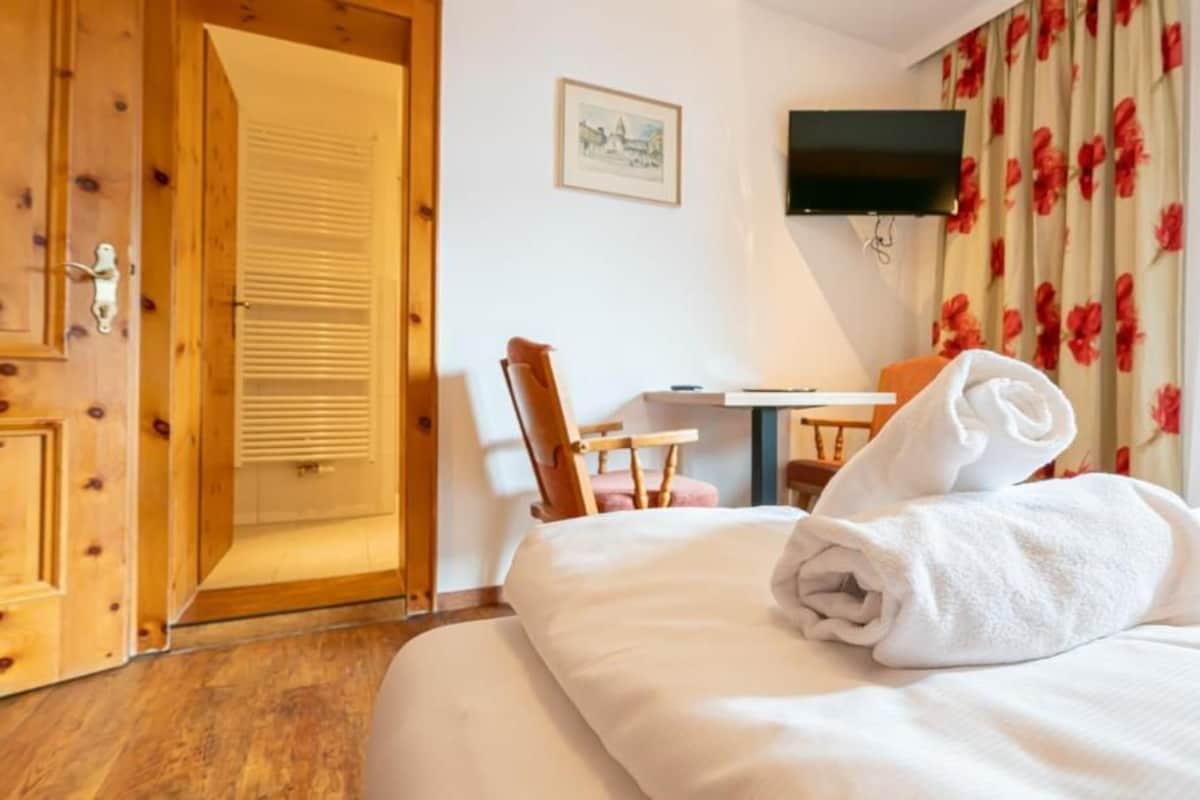 Una camera d'albergo con una porta in legno, un letto con un asciugamano arrotolato, una scrivania con una sedia e una televisione a parete.