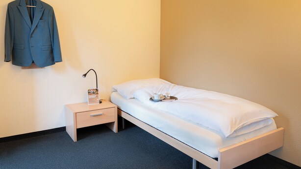 A single bed with white bedding, a bedside table with a lamp, a hanging coat, and a small nightstand.